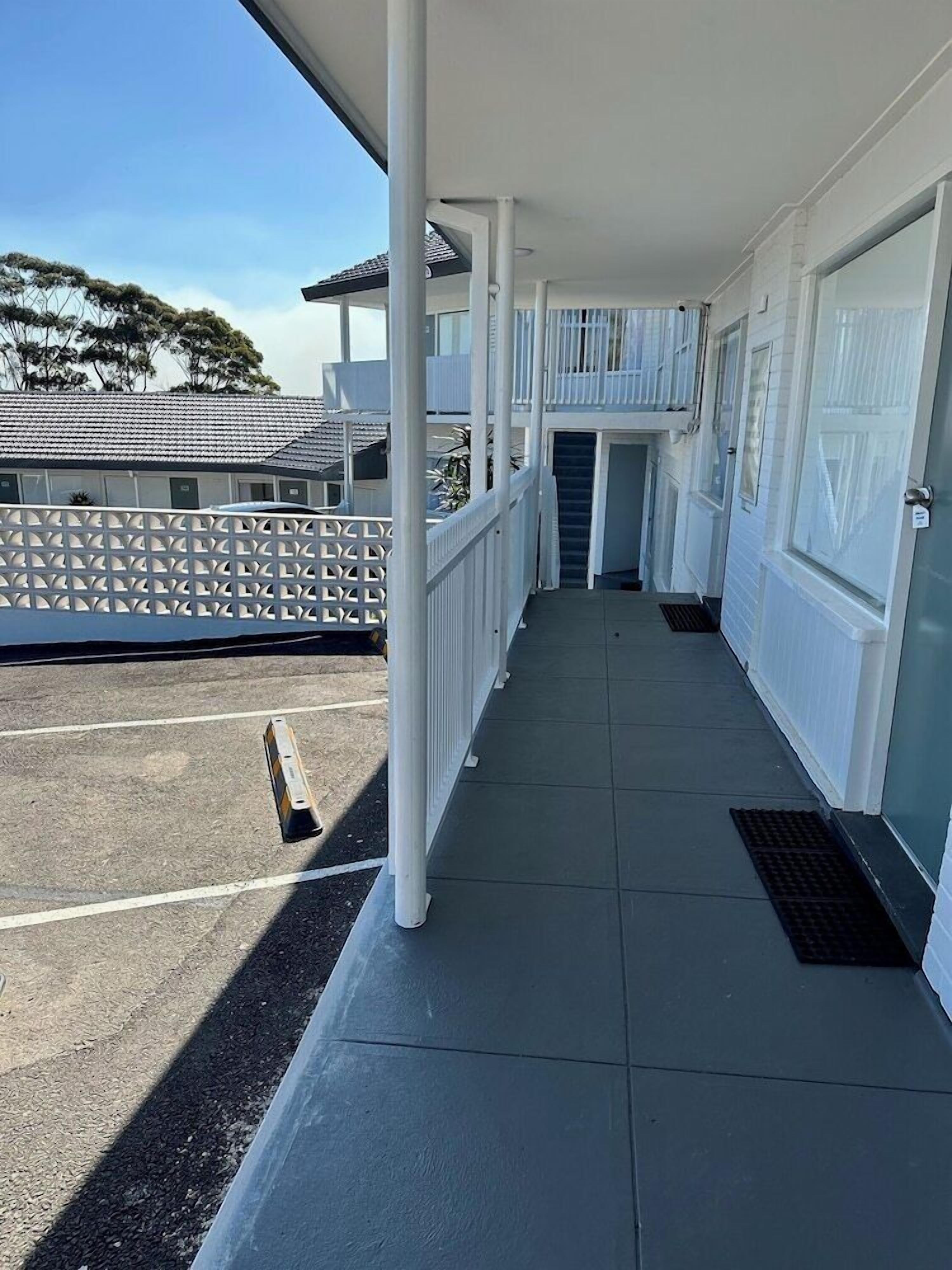 Un espace extérieur abrité, doté d’un sol carrelé, de rampes blanches et offrant une vue sur un bâtiment dont le toit est également recouvert de tuiles.