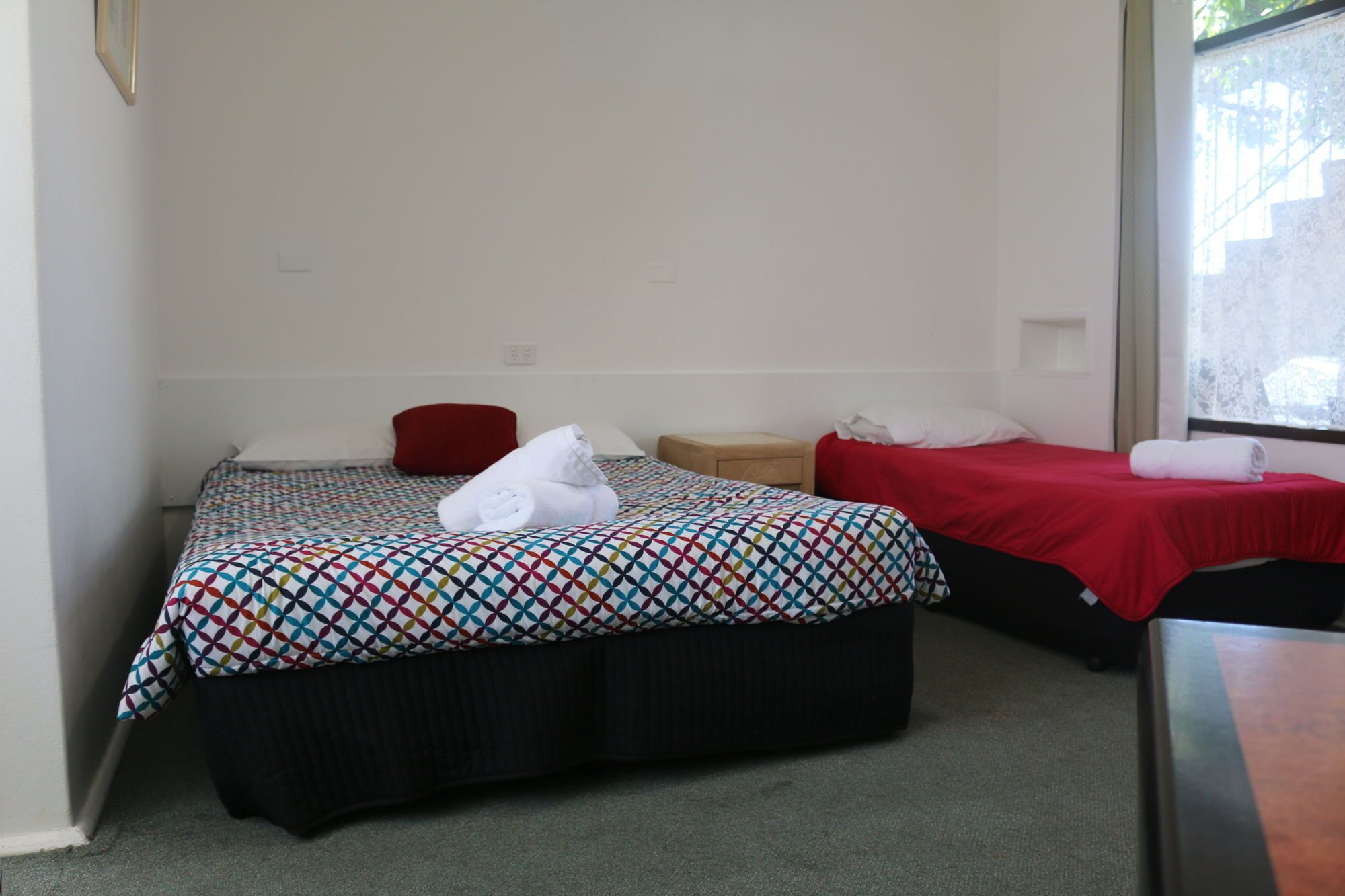 Habitación de hotel con dos camas, cada una con una colcha estampada, una almohada roja y una toalla blanca en una de ellas. Hay una mesita de noche de madera entre las camas.