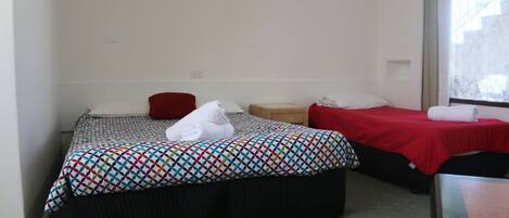 Habitación de hotel con dos camas, cada una con una colcha estampada, una almohada roja y una toalla blanca en una de ellas. Hay una mesita de noche de madera entre las camas.