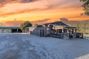 A rustic barn with a covered area, gravel ground, and a sunset sky. - The Homestead at GratiDude Ranch (Franklin)