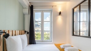 Ett hotellrum med en säng, ett fönster med utsikt över byggnader, en vägglampa och en handduksställning.