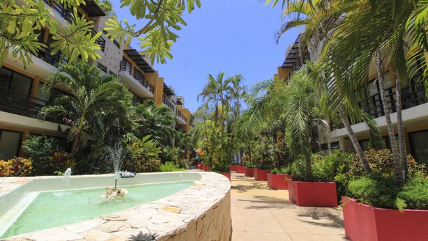 Un patio con una fuente central, rodeado de abundante vegetación y palmeras, con edificios de varios pisos a ambos lados.