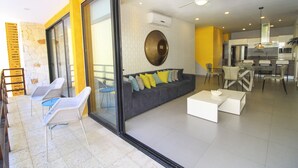 Un salón moderno con un sofá gris, sillas blancas y un ventanal de vidrio que da a una cocina.