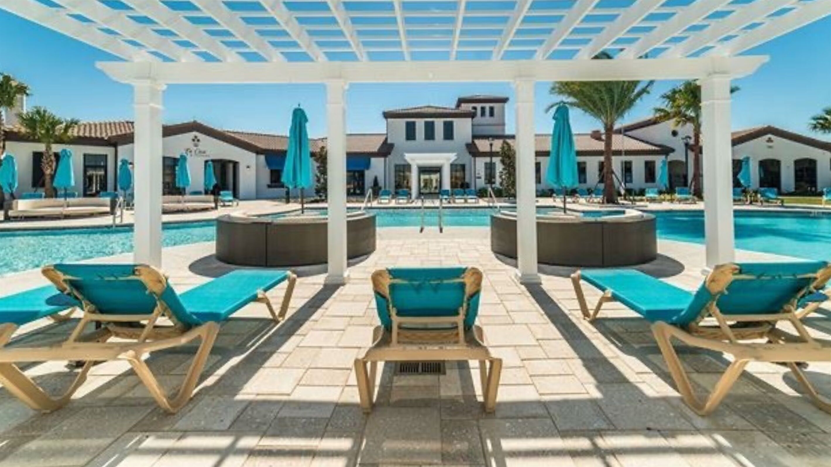 A pool area with lounge chairs and umbrellas, a covered seating area, and a building in the background.