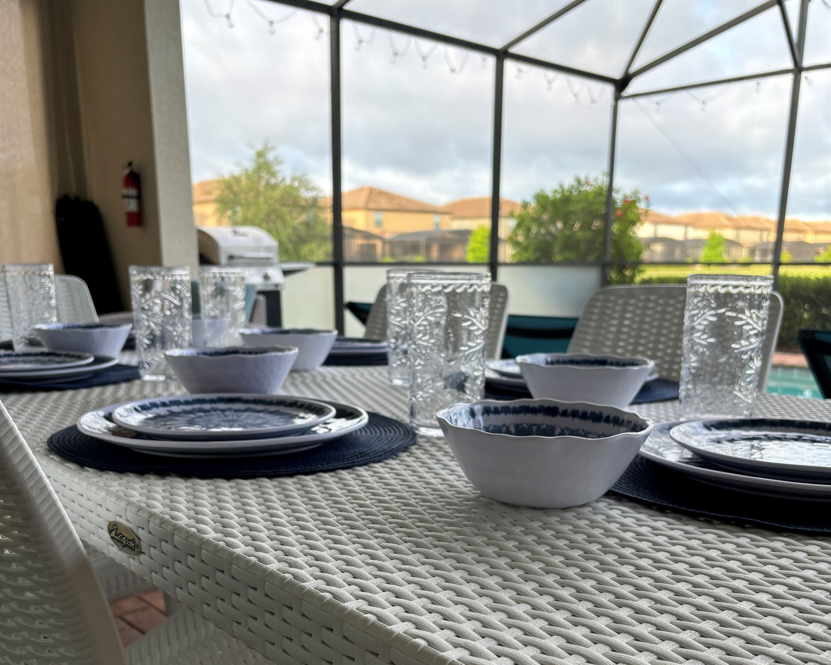 A table set with plates, bowls, and glasses, with a view of houses and trees through a glass enclosure.