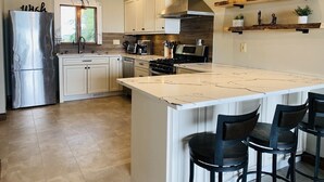 A modern kitchen with white cabinets, stainless steel appliances, a large island with bar stools, and wooden shelves with decorative items. - LAKE Escape Retreat - Onsite Beach Dock Waterfront Boat Ramp (Amherstburg)