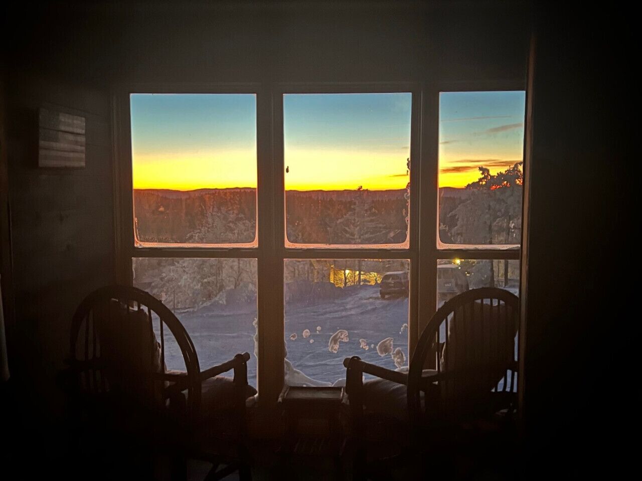 A room with a large window overlooking a snowy landscape during sunset. Two wooden chairs are positioned in front of the window.
