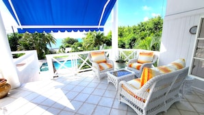 Une terrasse extérieure couverte, aménagée avec des meubles en osier, une piscine et une végétation luxuriante en arrière-plan.