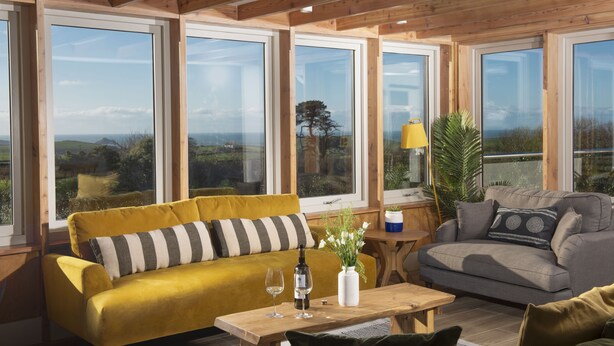 A spacious room with large windows, a yellow sofa, a grey sofa, and a wooden table with a wine bottle and glasses. - Westaway House in Trelights near Port Isaac (Port Isaac)