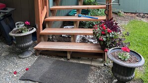 Una escalera de madera que lleva a una puerta, rodeada de plantas en macetas y un felpudo.