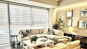 Een moderne woonkamer met een hoekbank, een salontafel en twee fauteuils. Er is een klein bijzettafeltje met een lamp, een potplant en een lijst met een foto aan de muur.