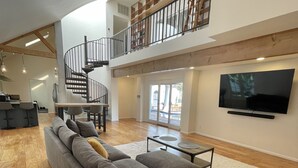 Ein geräumiges Wohnzimmer mit einem großen grauen Ecksofa, einer Holztreppe und einem an der Wand befestigten Flachbildfernseher.
