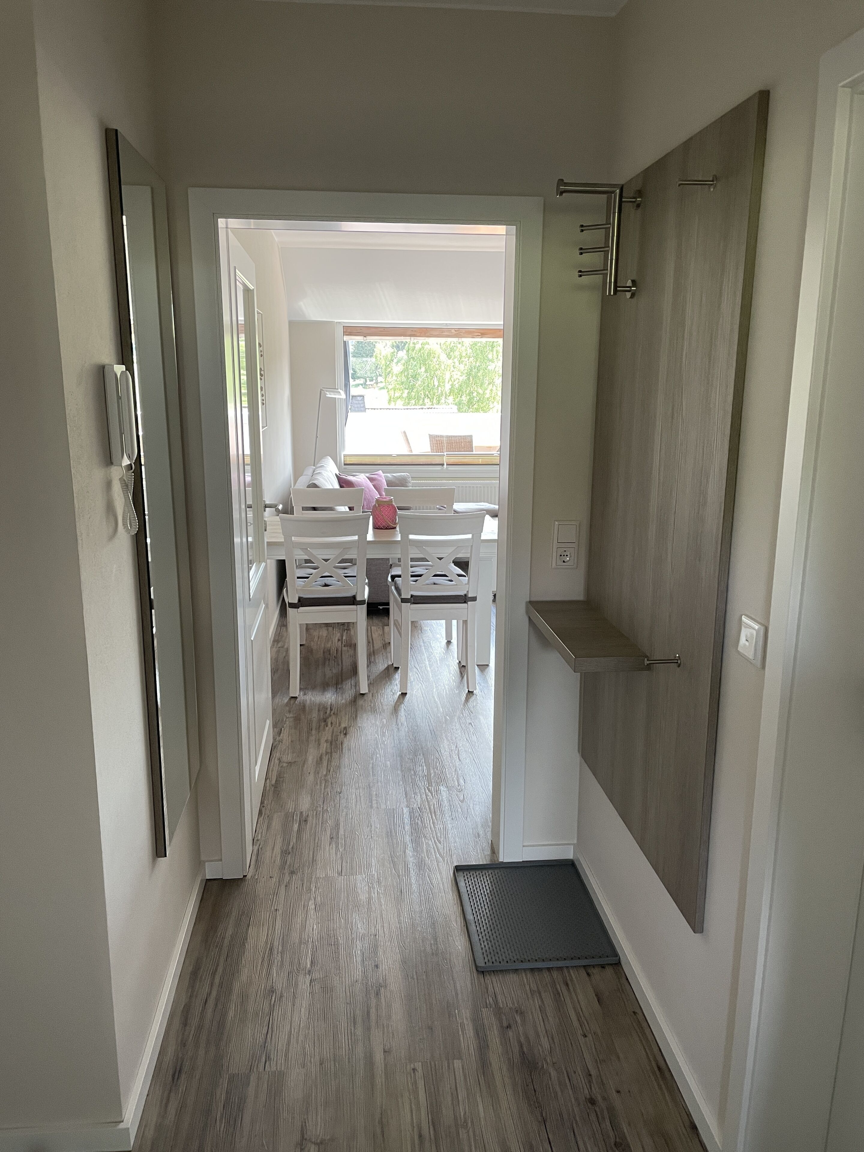 Un couloir étroit, doté d’un plancher en bois, mène à une pièce avec une fenêtre, une chaise blanche et une petite table.