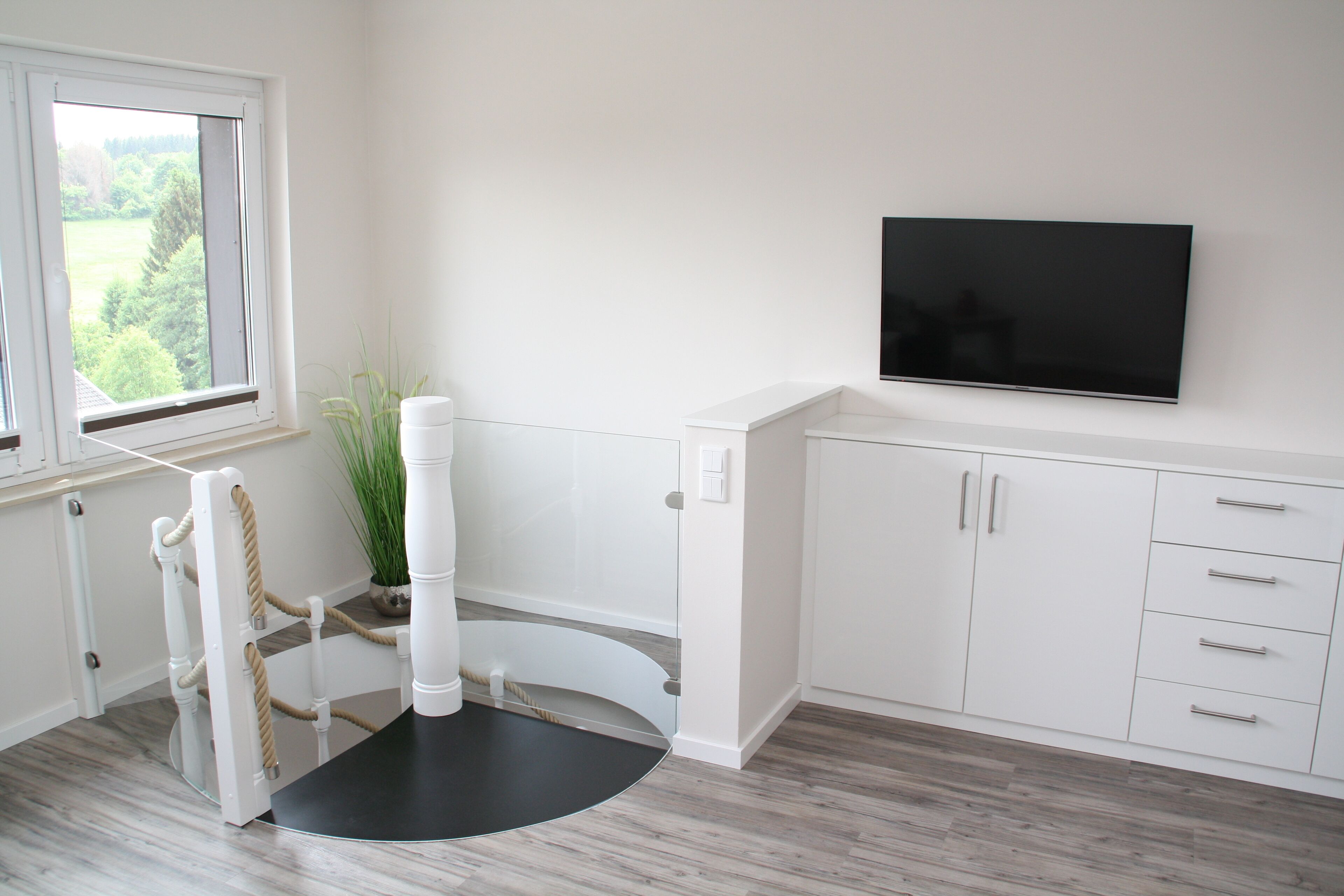 Une chambre moderne avec une grande fenêtre, un téléviseur à écran plat fixé au mur, des meubles blancs et une plante en pot.