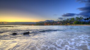 Escena de playa con olas, un atardecer y una zona turística con edificios y palmeras.