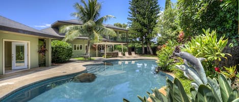 Un área de piscina con diseño curvo, rodeada de plantas tropicales y un gran árbol.