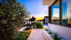 Una casa moderna con un sendero que lleva a una piscina, rodeada de vegetación y una vista despejada del atardecer.