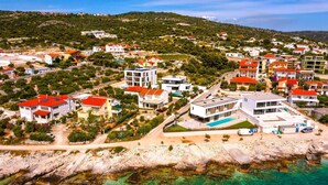 Vista aérea de una zona residencial costera con casas, mar azul cristalino y un paisaje montañoso al fondo.