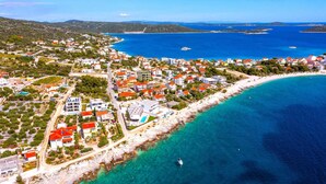 Vista aérea de un pueblo costero con playa, aguas azules cristalinas y edificios con techos rojos.