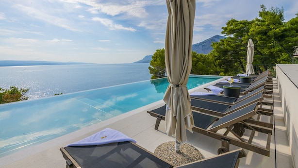 An infinity pool with lounge chairs and umbrellas, overlooking a body of water and a mountainous landscape. - Villa Azul Grande with Heated Pool (Brela)