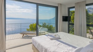 A bedroom with a large bed, a balcony with a view of the sea, and a TV mounted on the wall. - Villa Azul Grande with Heated Pool (Brela)