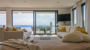 A modern living room with two sofas, a coffee table, and a TV. The room has large windows offering a view of the sea. - Villa Azul Grande with Heated Pool (Brela)
