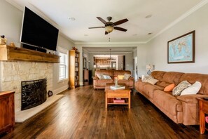 A spacious living room with hardwood floors, a fireplace, a large flat-screen TV, a ceiling fan, and a brown leather sofa.