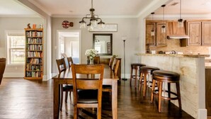 Un comedor con mesa y sillas de madera, una estantería y una cocina con armarios de madera y luces colgantes.