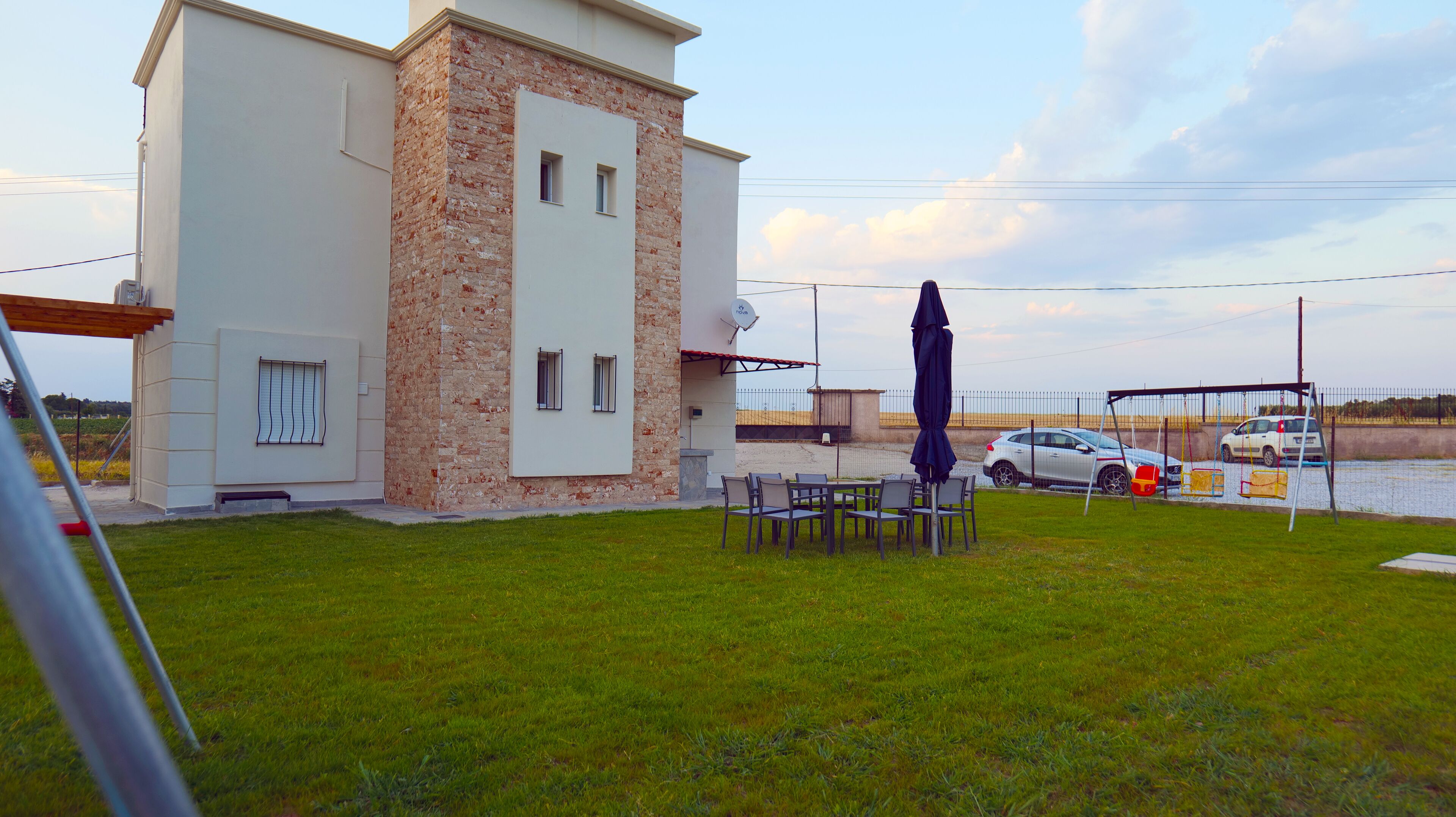 A modern building with a stone facade, a patio with chairs and an umbrella, and a car parked by a water body.