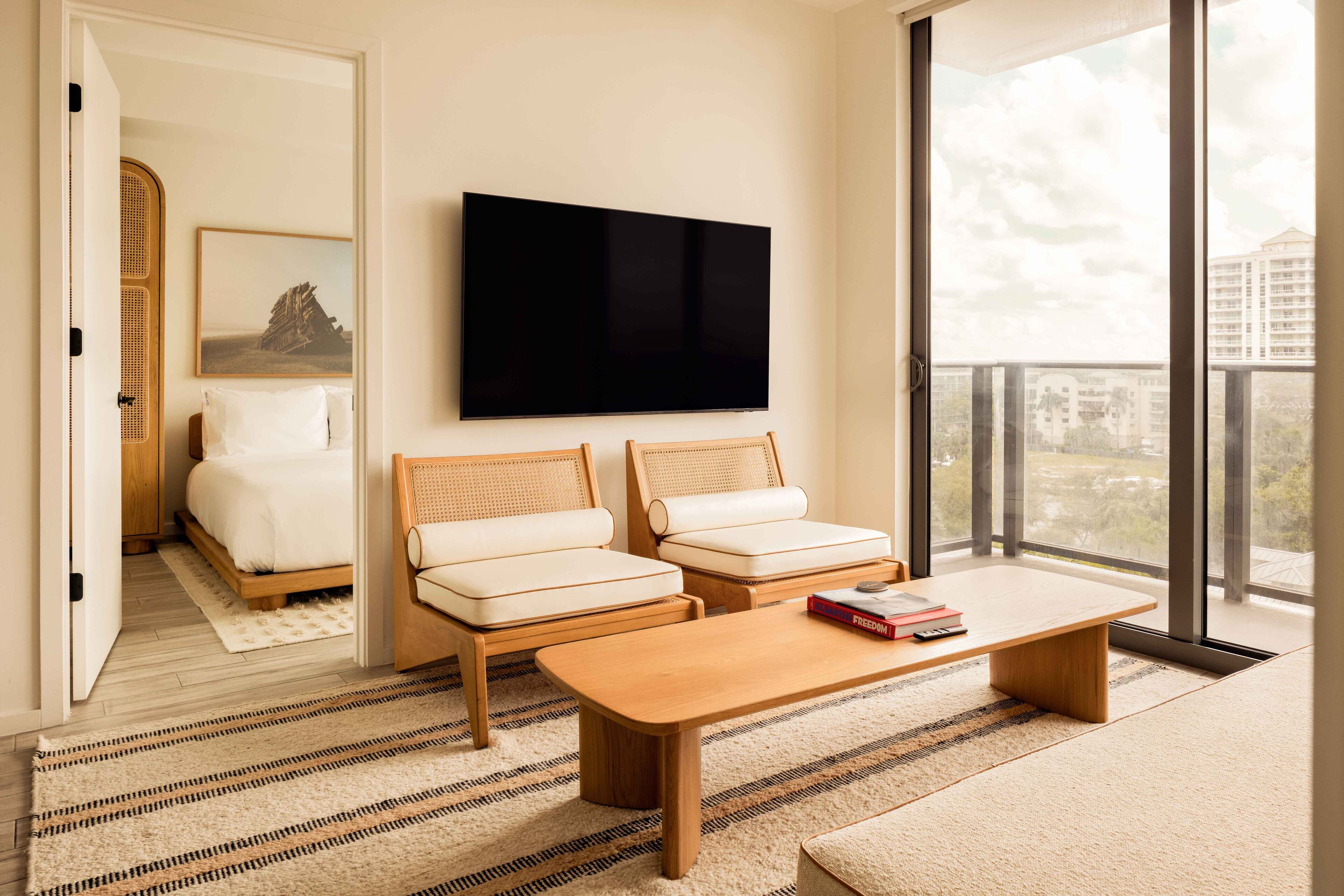 A modern hotel room with a large bed, two sofas, a coffee table, a flat-screen TV, and a large window offering a city view.