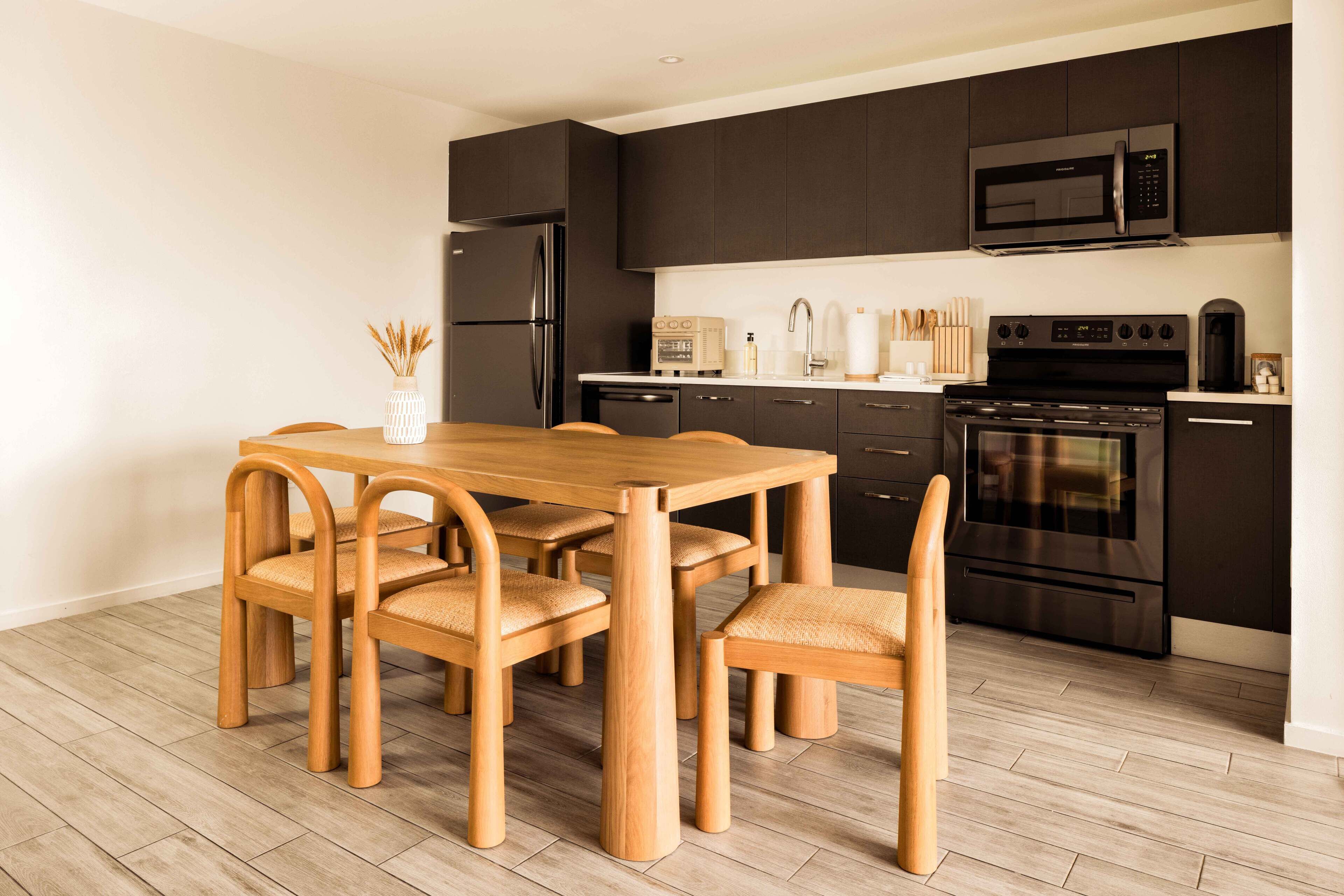 Una cocina moderna con armarios oscuros, un refrigerador de acero inoxidable, un microondas, un horno y una mesa de comedor con sillas de madera.