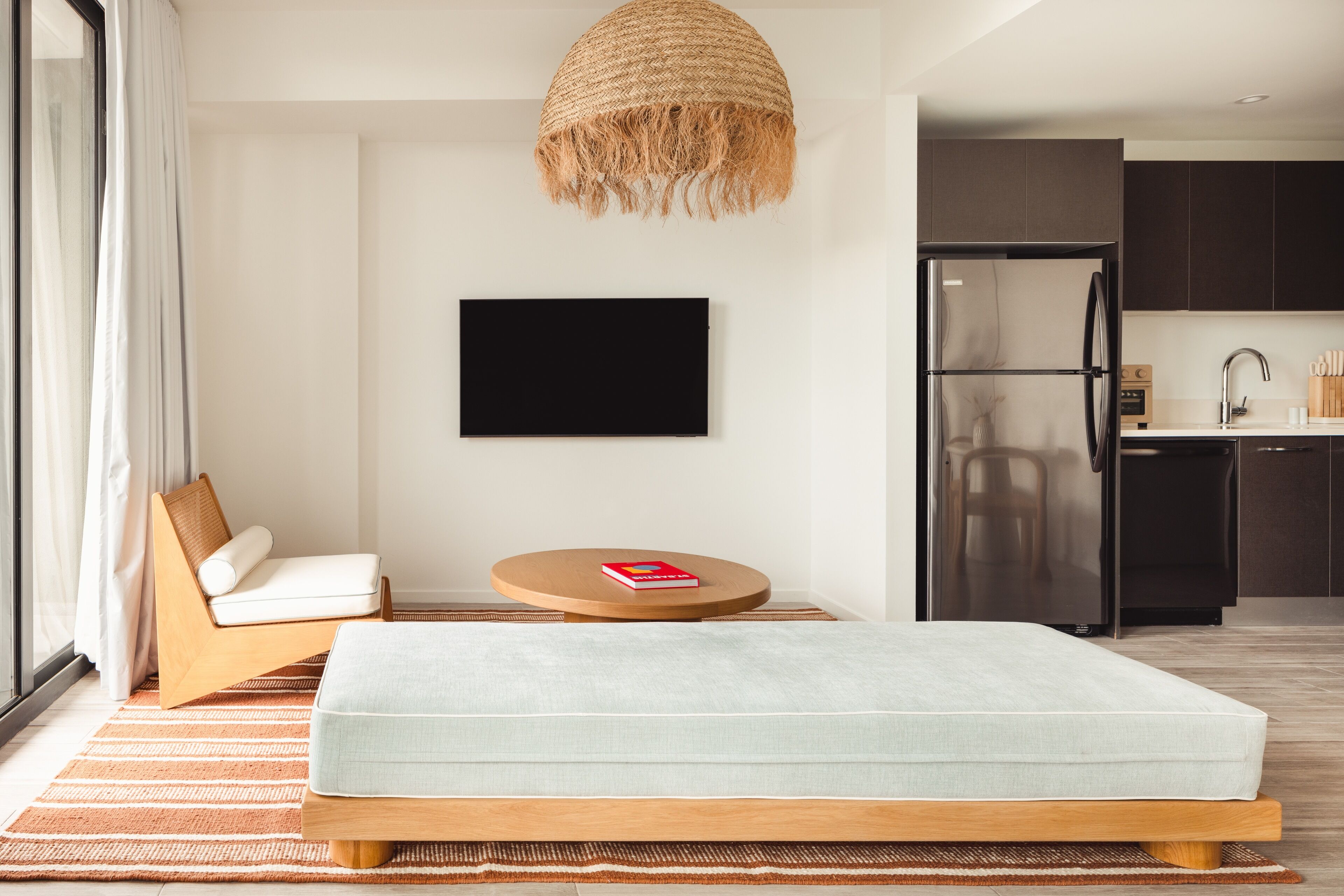 A modern bedroom with a bed, a TV, a chair, and a kitchen area in the background.