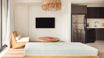 A modern bedroom with a bed, a TV, a chair, and a kitchen area in the background.