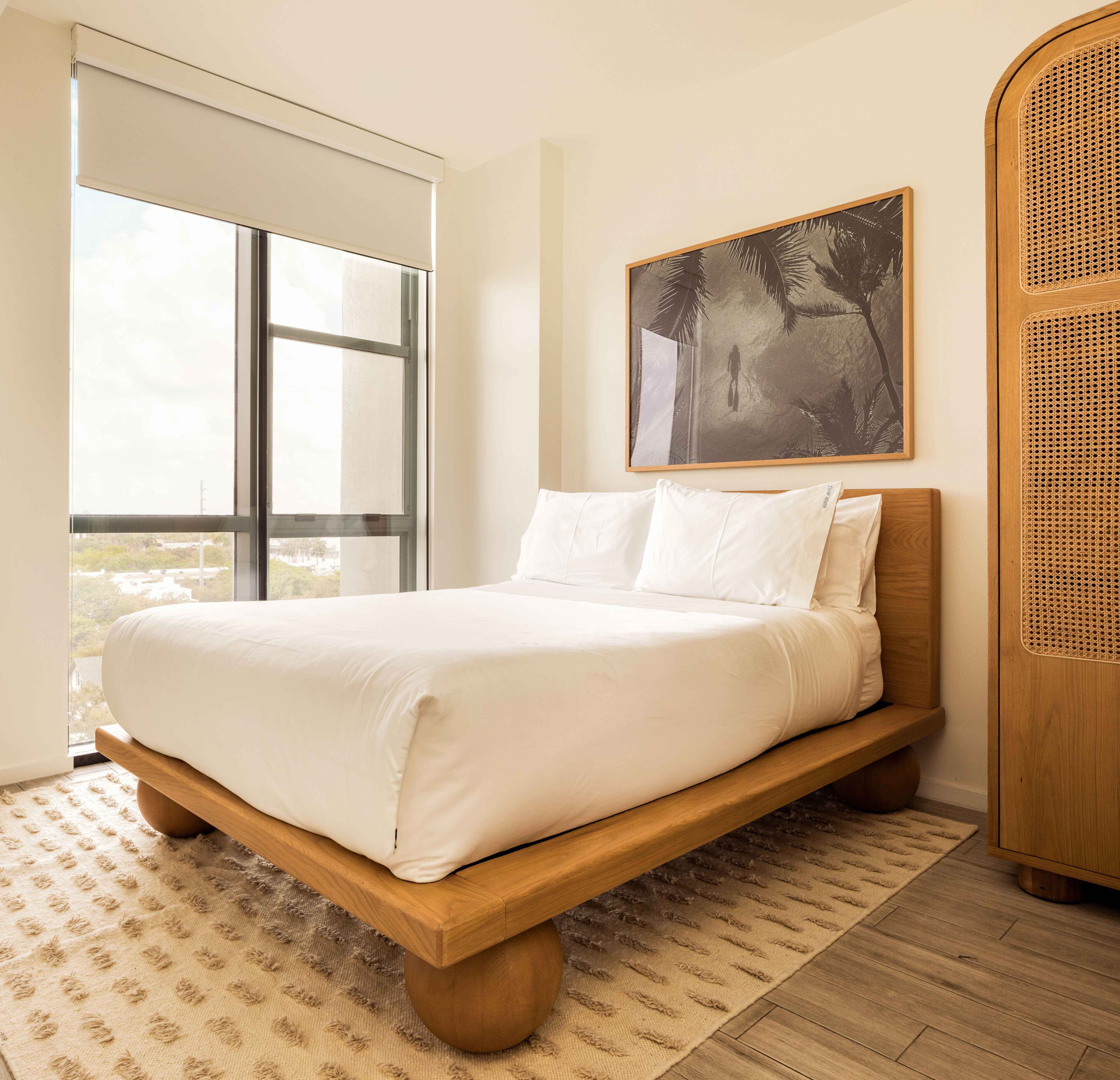 A bedroom with a large window, a bed with white bedding, a rattan headboard, and a framed picture on the wall.
