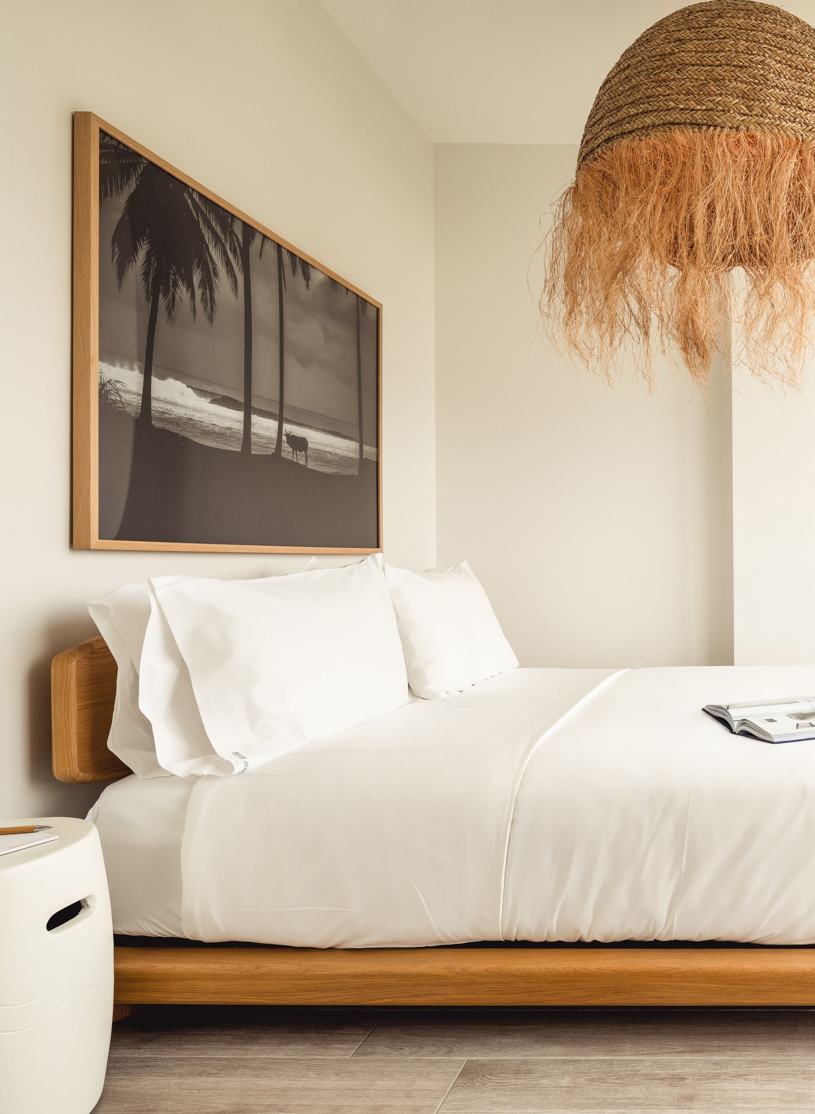 A bedroom with a bed, pillows, a wooden headboard, a bedside table, a white lamp, and a framed picture on the wall.