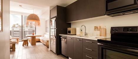 A modern kitchen with dark cabinets, a built-in microwave, and an oven. There is a dining area with a table and chairs, and a large window offering a view of the city.