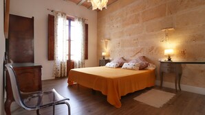 Un dormitori amb un llit, dues taules de nit amb làmpades, una cadira, un escriptori de fusta, una finestra amb cortines i una paret de pedra.