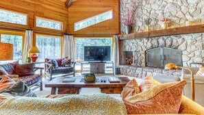 A cozy cabin interior with a stone fireplace, wooden beams, and a large window overlooking a forest. - Pools, Private Hot Tub & Pool Table. Upscale Lodge (Sisters)