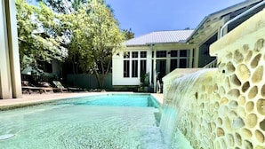 Una piscina cristalina con una cascada, rodeada de sillones de descanso y un edificio con amplios ventanales.