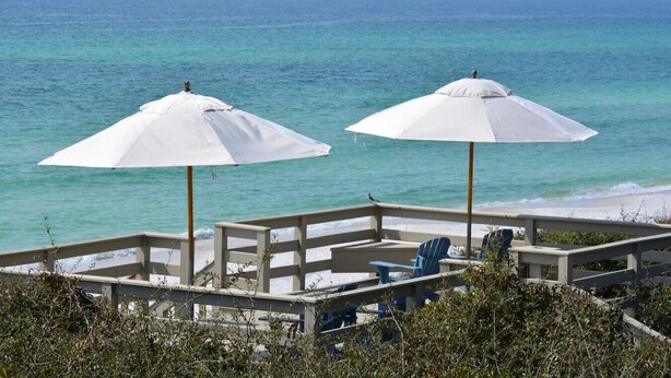 Zona de estar junto a la playa, con dos sombrillas blancas y algunas sillas.