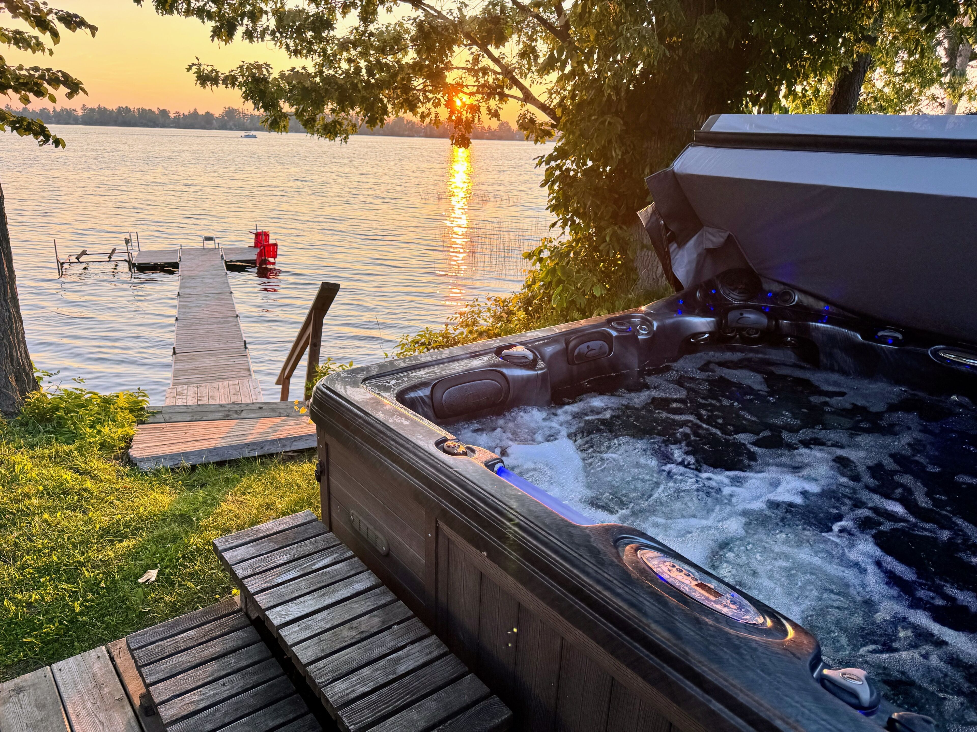 Een hottub met een houten terras dat uitkomt op een steiger, met uitzicht op een rustig meer bij zonsondergang.