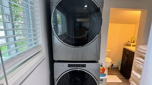Een wasmachine en droger boven elkaar in een wasserij, met een raam, een deur naar een andere ruimte en een witte handdoekhouder.
