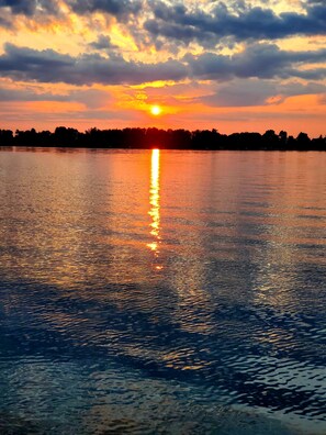 Un coucher de soleil sur une étendue d’eau calme, avec la silhouette d’arbres en arrière-plan.