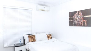 Un dormitorio con cama, ropa de cama blanca, una mesita de noche, una botella de agua, un control remoto, aire acondicionado instalado en la pared y una obra de arte abstracta.