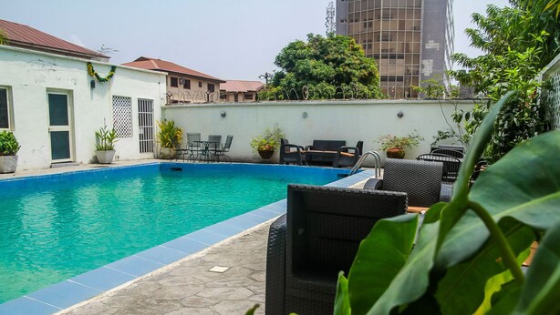 Una piscina con sillas de descanso y plantas en macetas, rodeada de edificios y follaje.