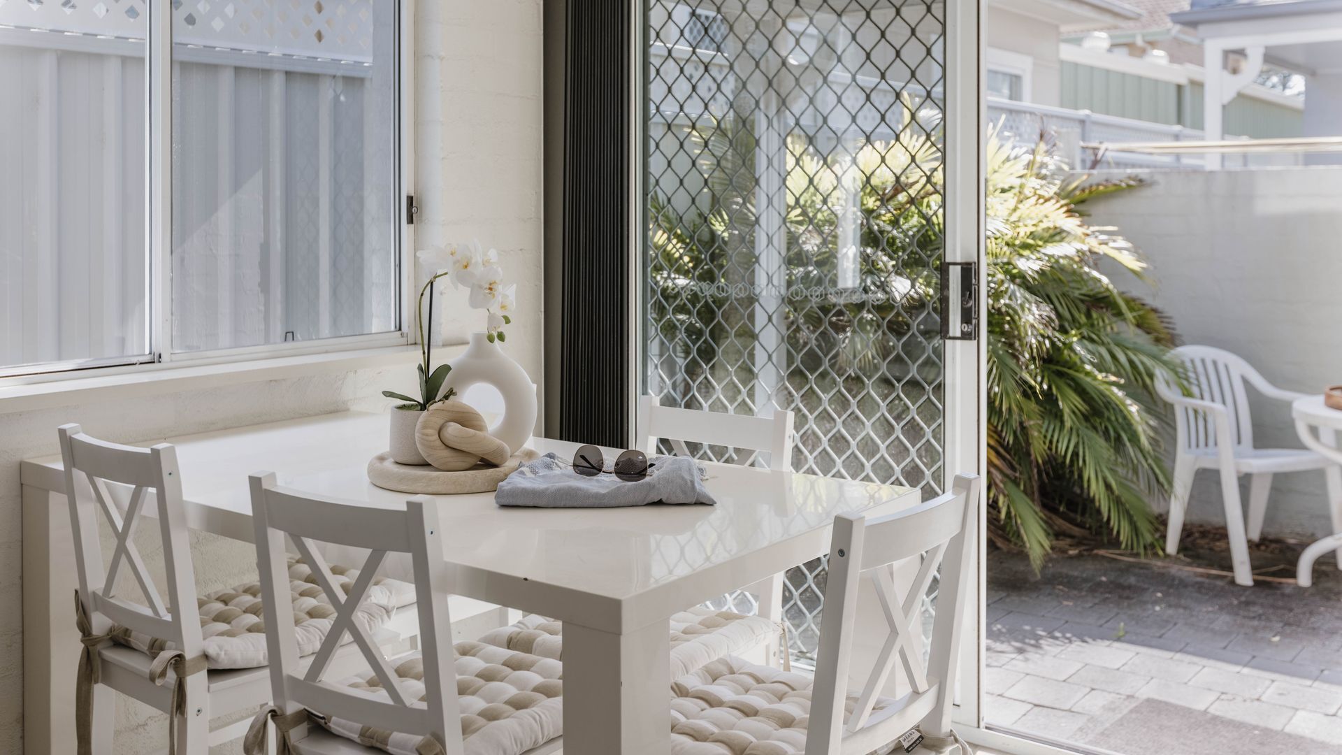 A white dining table with four chairs, a vase with flowers, and a pair of sunglasses on a cushion.