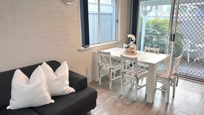 Ein Zimmer mit einer schwarzen Couch, weißen Kissen, einem weißen Esstisch mit Stühlen und Blick durch eine Glastür auf einen Außenbereich mit Zaun und Grünpflanzen.