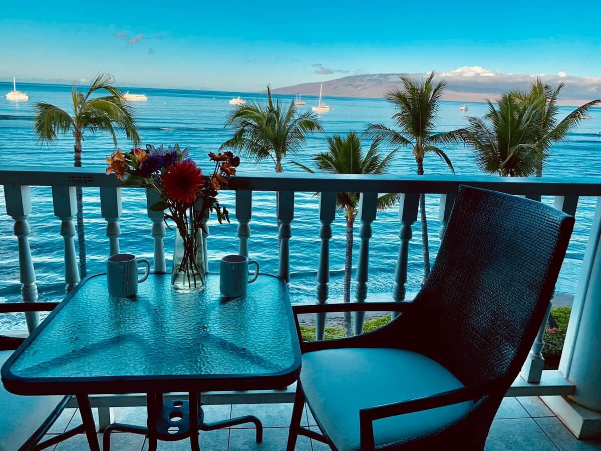 A balcony with a table and chair, overlooking a clear blue sea with boats and palm trees.