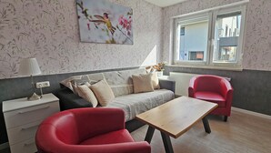 A cozy living room with a sofa, two red chairs, a wooden coffee table, a lamp, and a window with a view of buildings. - Apartment Fulda 65m² up to 6 pers. (Fulda)