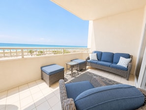A balcony with a wicker sofa, a small table, and a cushioned bench, overlooking a beach.